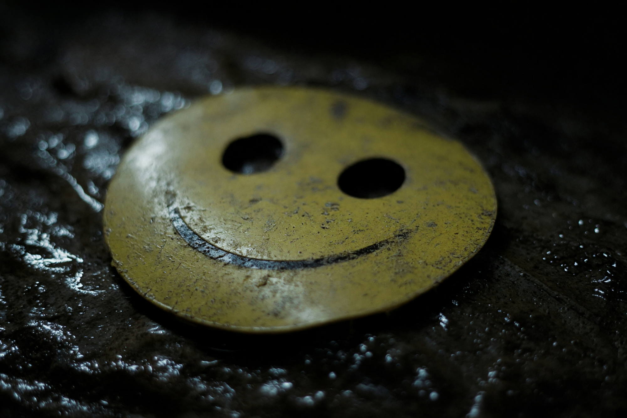 Macro photograph of a scuffed yellow plastic smiley mask lying on a wet, dark, glistening surface.