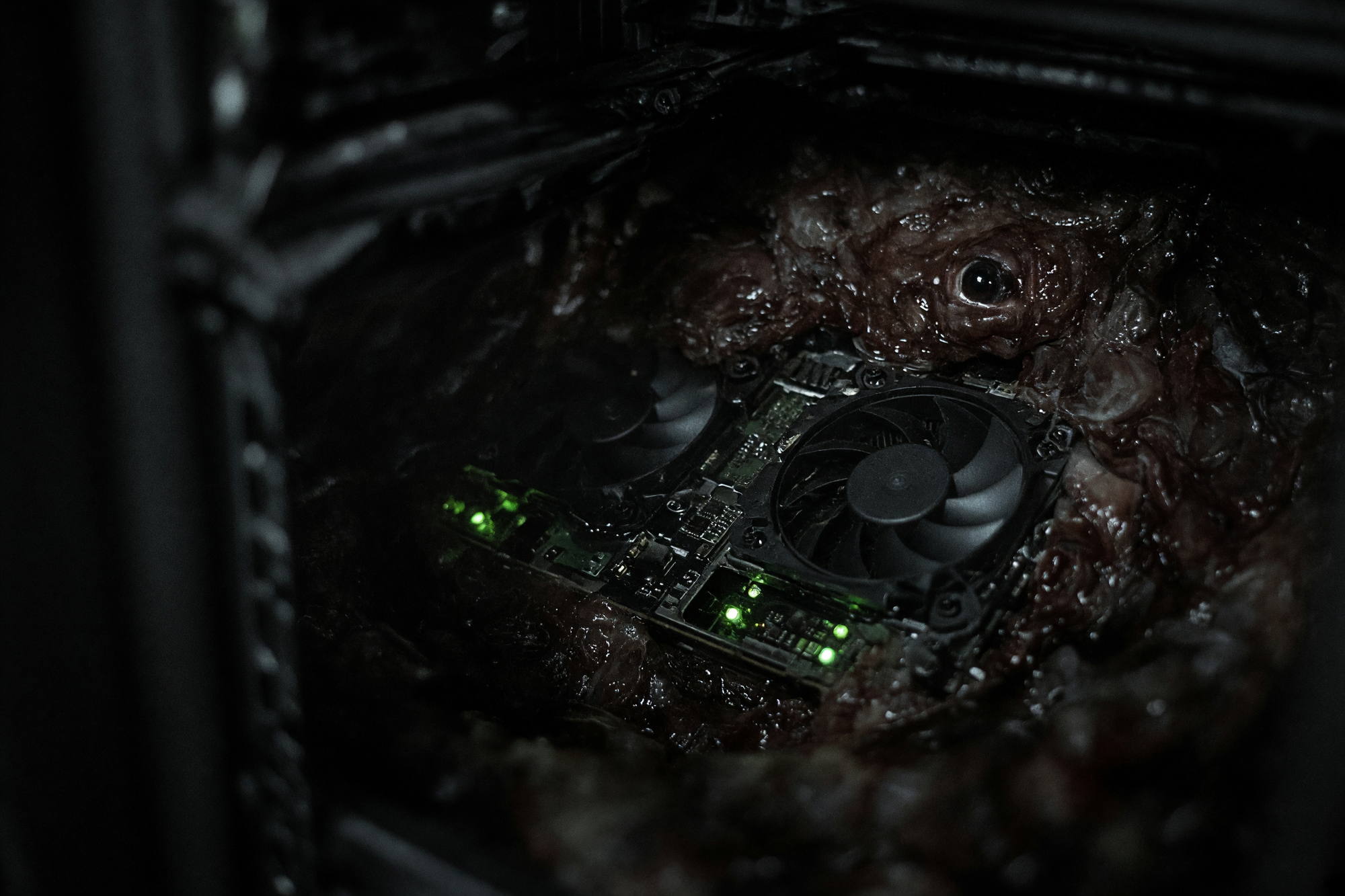 A graphics card inside an open rack, green status lights on, with dark tissue and a single eye grown around it.